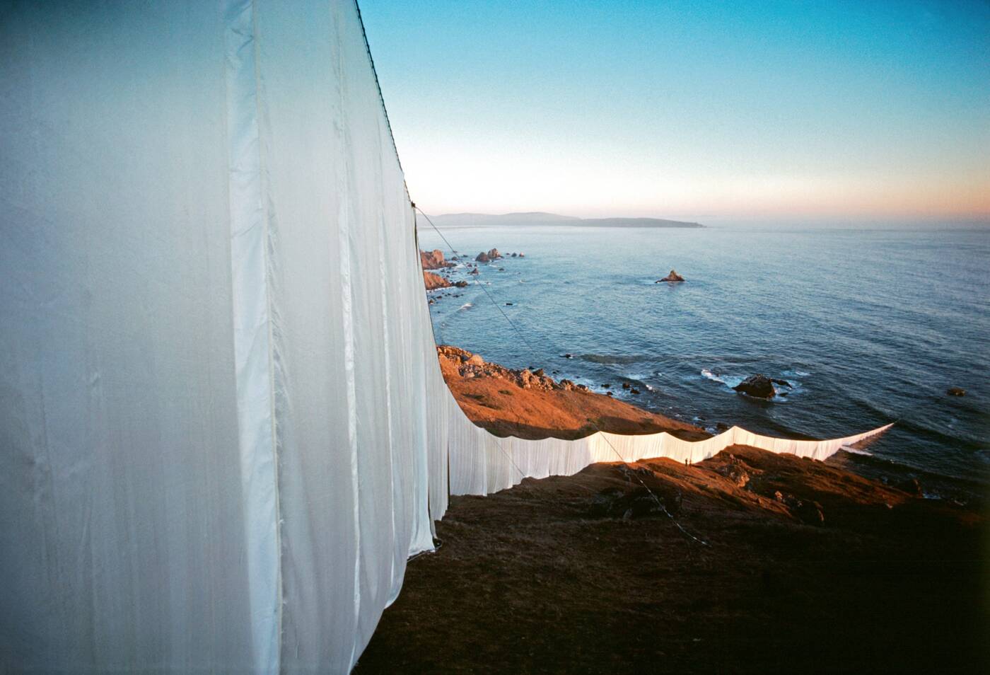 Running Fence desce por uma encosta rochosa e alcança o oceano Pacífico ao entardecer.