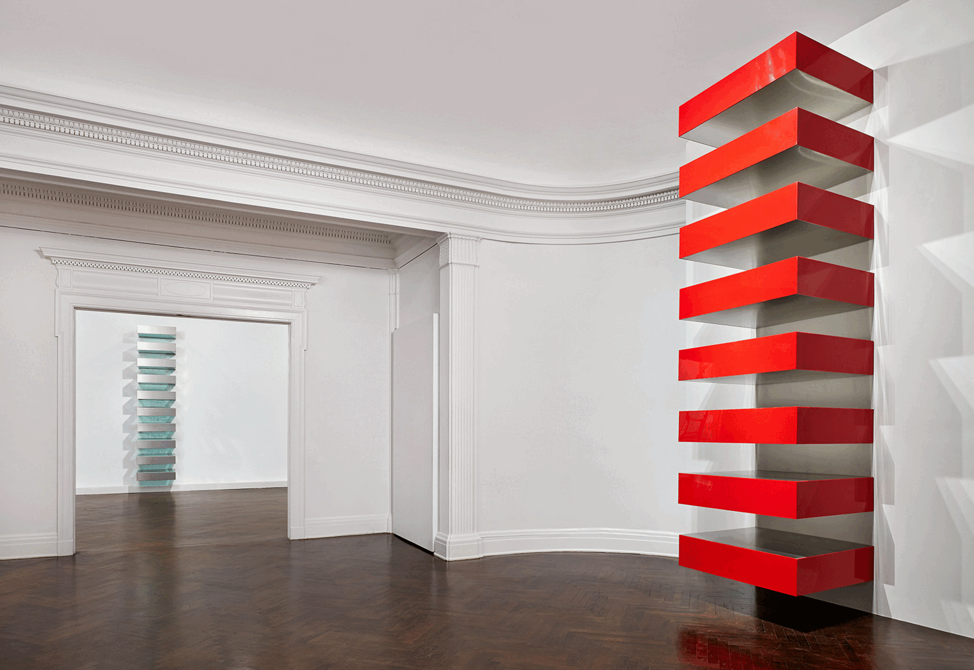 Duas Stacks de Donald Judd, uma vermelha em primeiro plano e outra verde ao fundo, instaladas em salas interligadas.