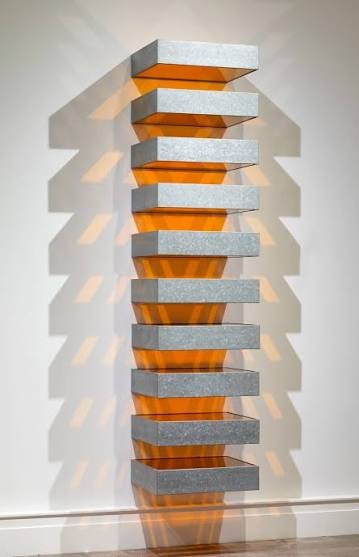 Stack de Donald Judd com módulos metálicos e superfícies alaranjadas translúcidas, projetando sombras sobre a parede.