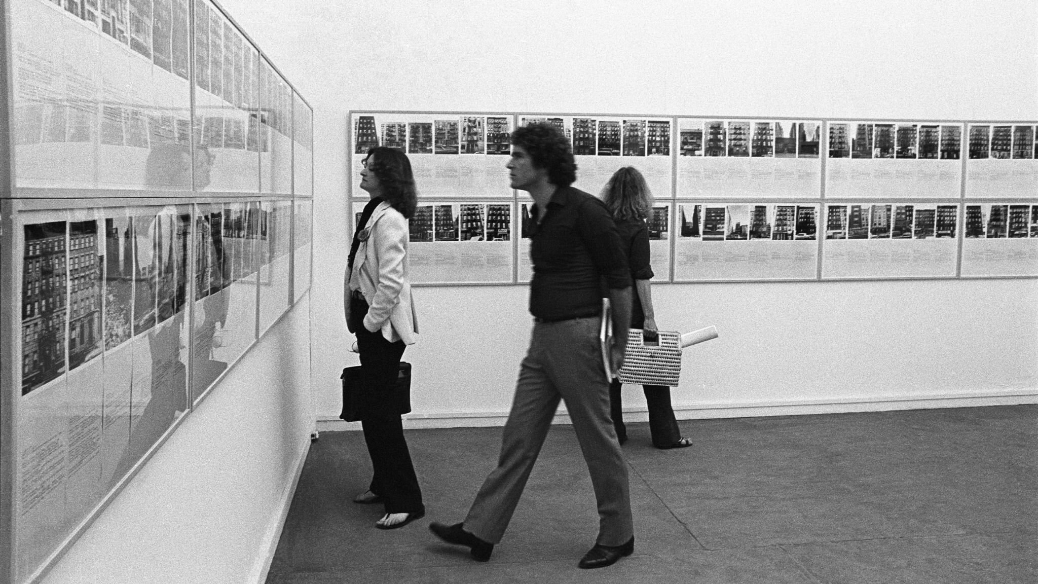 Visitantes observam a extensa instalação de Shapolsky et al. em uma galeria, com fotografias e documentos ocupando diferentes paredes.