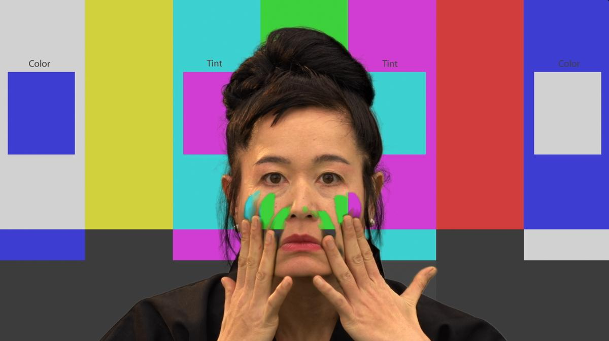 Hito Steyerl aplica manchas coloridas no rosto diante de barras de cor e campos de chroma key.
