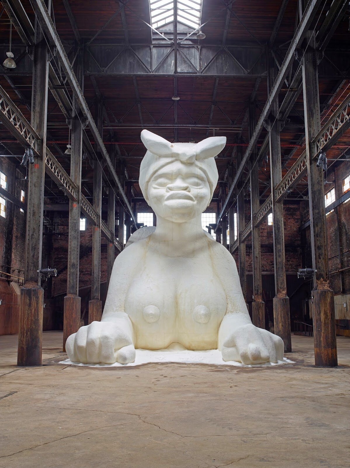 Escultura monumental de Kara Walker em forma de esfinge com feições de uma mulher negra, revestida de açúcar branco, no interior da antiga refinaria Domino.