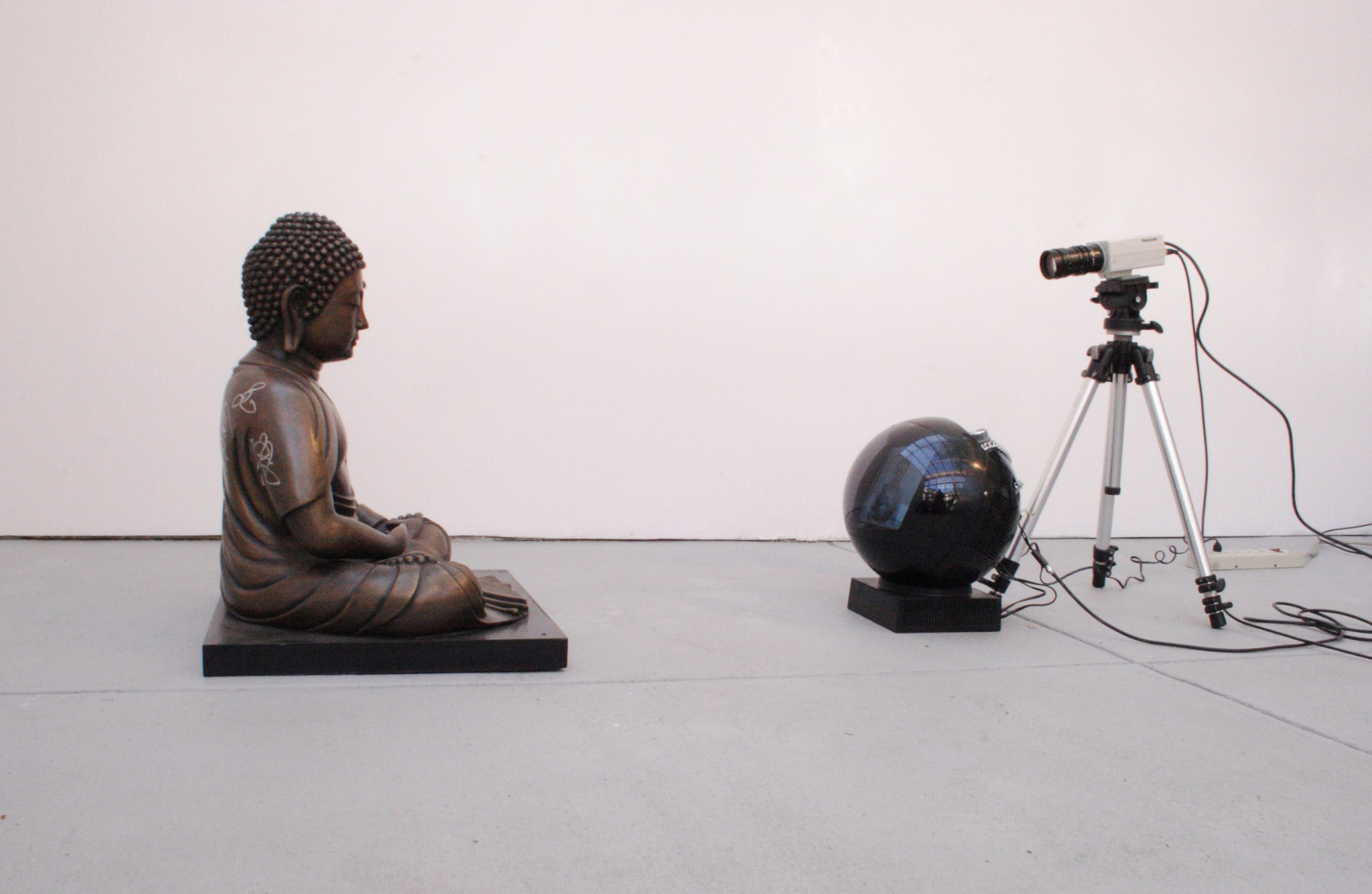 Vista lateral de uma versão de TV Buddha com escultura sentada, câmera sobre tripé e pequeno televisor esférico conectados por cabos.