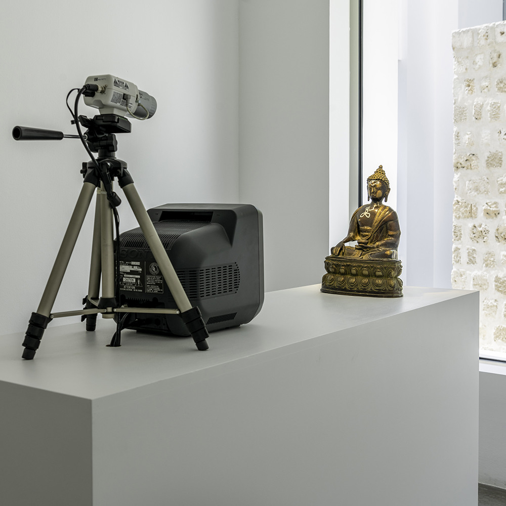 Vista posterior do equipamento de TV Buddha com câmera sobre tripé, monitor de tubo e pequena escultura dourada dispostos sobre um pedestal.