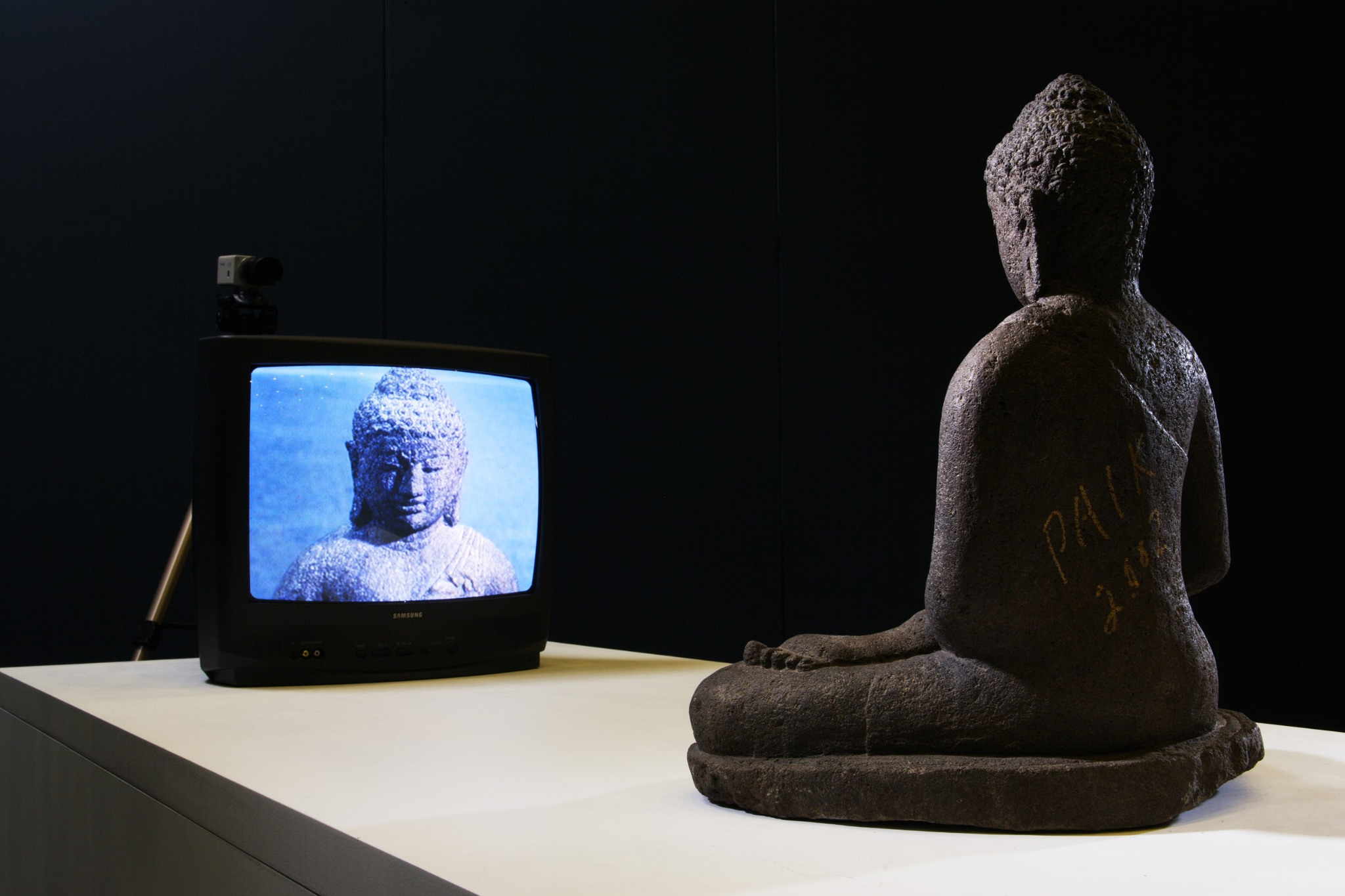 Instalação de TV Buddha, de Nam June Paik: uma escultura de Buda está sentada diante de um televisor que transmite sua imagem captada ao vivo por uma câmera.