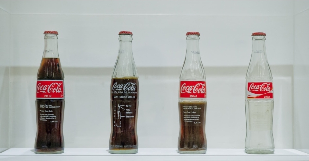 Quatro garrafas retornáveis de Coca-Cola, cheias e vazias, com mensagens críticas impressas sobre o vidro