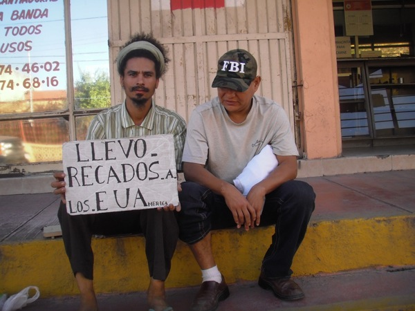Paulo Nazareth sentado ao lado de outro homem, segurando uma placa com a frase Llevo recados a los E.U.A.
