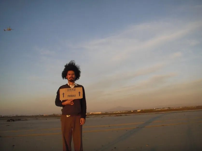 Paulo Nazareth em uma pista de aeroporto segurando uma caixa marcada com a palavra Fragile
