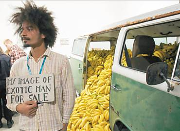 Paulo Nazareth ao lado de uma van carregada de bananas, segurando a placa My image of exotic man for sale