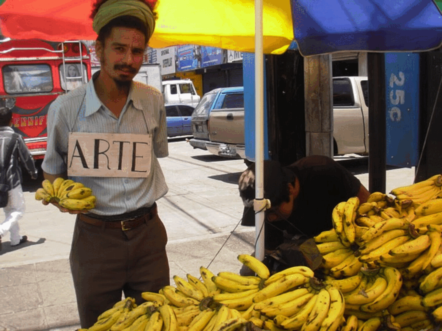 Paulo Nazareth em uma banca de bananas segurando frutas e uma placa com a palavra Arte